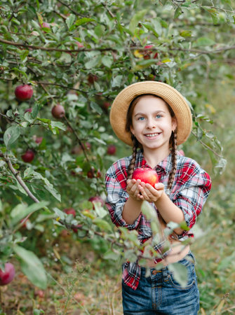 Cheerful girl stands on background an apple orchard with red apples, tastes, enjoys snacks, vitamins, organic fruits. She is dressed in checkered shirt and hat. Family picnic, Autumn apple harvest.の写真素材