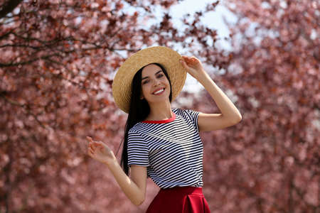 Pretty young woman with straw hat near beautiful blossoming trees outdoors. Stylish spring lookの写真素材