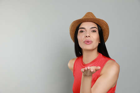 Beautiful young woman with straw hat blowing kiss on light grey background. Space for text