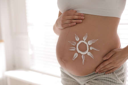Young pregnant woman with sun protection cream on belly indoors, closeup. Space for textの写真素材