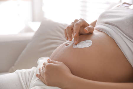 Young pregnant woman applying cosmetic product on belly indoors, closeupの写真素材