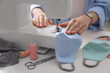 Woman making cloth mask with sewing machine at white table, closeup