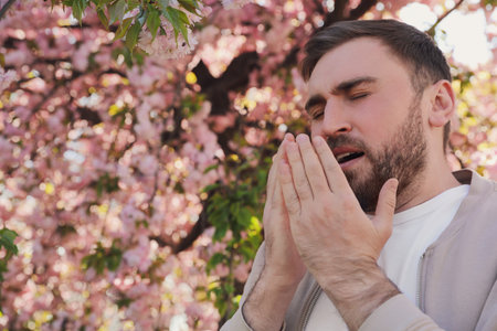 Man suffering from seasonal pollen allergy near blossoming tree outdoors