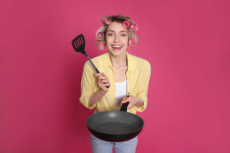 Young housewife with frying pan and spatula on pink backgroundの写真素材