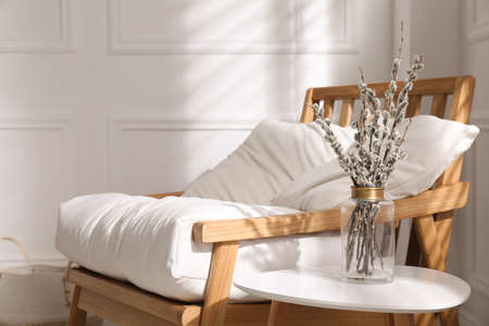 Glass vase with pussy willow tree branches on table near armchair indoors, space for textの写真素材
