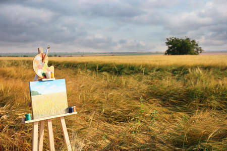 Wooden easel with beautiful picture and painting equipment in field. Space for textの写真素材