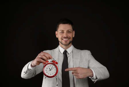 Happy businessman pointing on alarm clock against black background. Time managementの写真素材