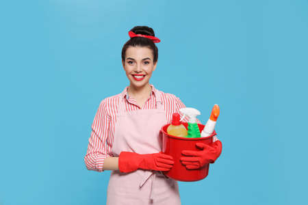 Young housewife holding bucket with cleaning supplies on light blue backgroundの写真素材