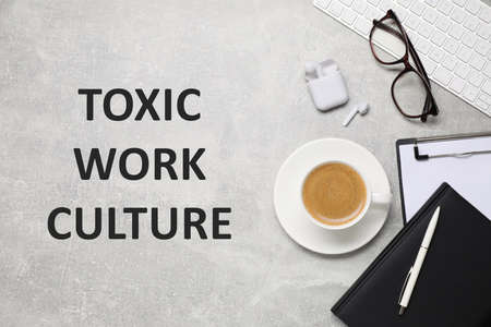 Cup of coffee, computer keyboard, stationery and text TOXIC WORK ENVIRONMENT on gray table, flat lay