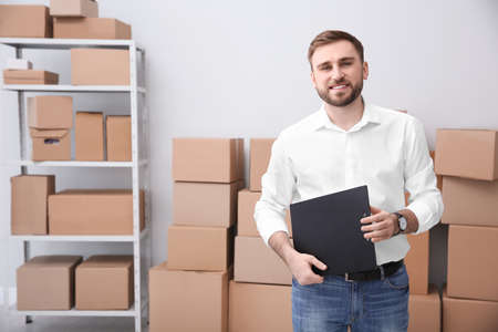 Young businessman with clipboard near cardboard boxes at warehouse