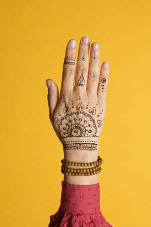 Woman with beautiful henna tattoo on hand against yellow background, closeup. Traditional mehndi