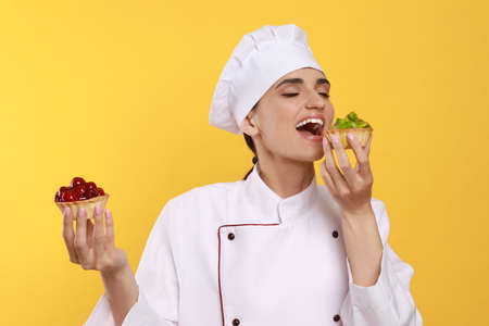Professional confectioner in uniform with delicious tartlets on yellow backgroundの写真素材