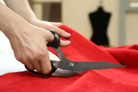 Seamstress cutting red fabric with scissors at workplace, closeup