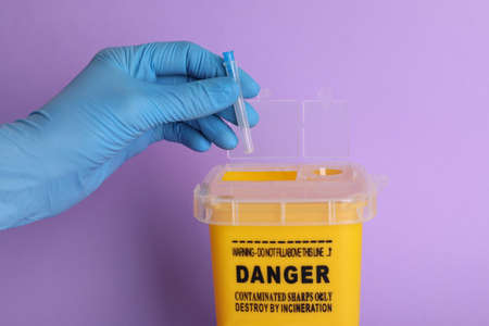 Doctor throwing used syringe needle into sharps container on violet background, closeup