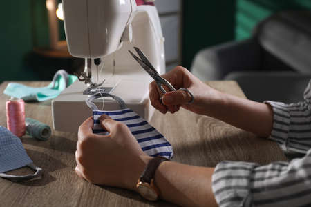 Woman sewing cloth protective mask with machine and scissors at table indoors, closeup