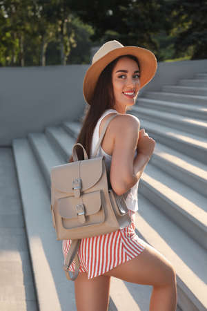 Beautiful young woman with stylish backpack and hat on stairs outdoorsの写真素材