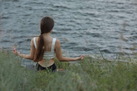 Teenage girl meditating near river, back view. Space for text