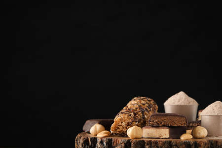 Different tasty energy bars, nuts and protein powder on wooden stump against black background, space...