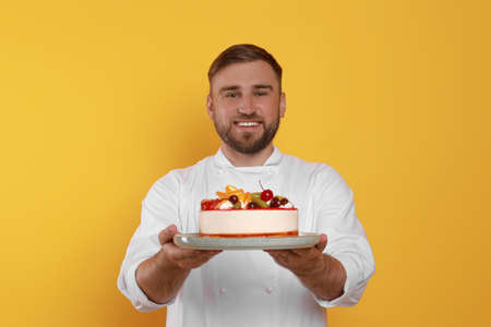 Happy professional confectioner in uniform holding delicious cake on yellow backgroundの写真素材