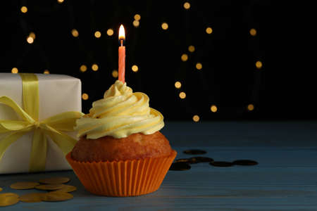 Delicious birthday cupcake with candle near gift box on wooden table against blurred festive lights, space for textの写真素材