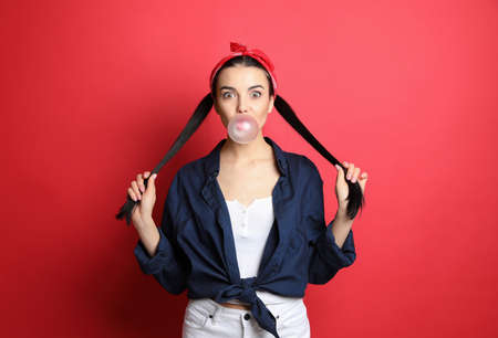 Fashionable young woman in pin up outfit blowing bubblegum on red background