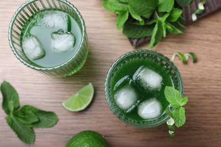 Delicious mint liqueur with green leaves and ice cubes on wooden table, flat lay