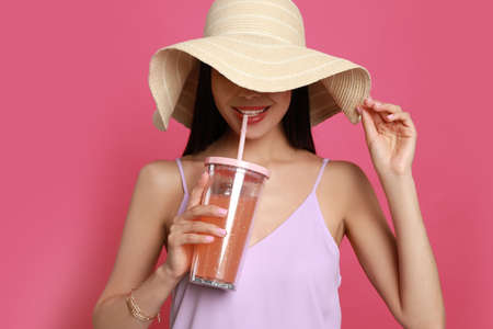 Beautiful young woman with straw hat and glass of refreshing drink on pink background
