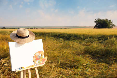 Wooden easel with blank canvas, painting equipment and hat in field. Space for textの写真素材