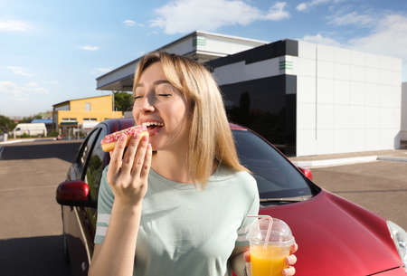 Beautiful young woman with juice eating donut near car at gas stationの写真素材