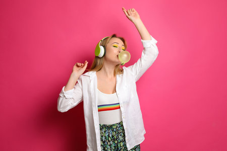 Fashionable young woman with bright makeup and headphones blowing bubblegum on pink backgroundの写真素材