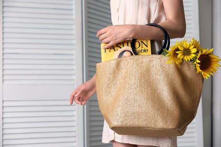 Woman holding beach bag with magazine and beautiful bouquet of sunflowers indoors, closeupの写真素材