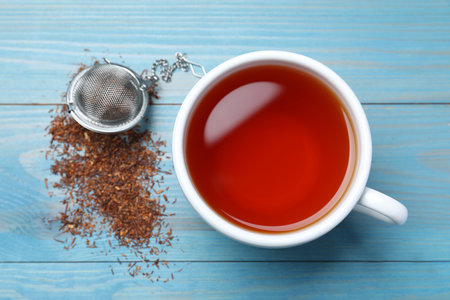 Ceramic cup of aromatic rooibos tea, infuser and scattered dry leaves on light blue wooden table, flat layの写真素材