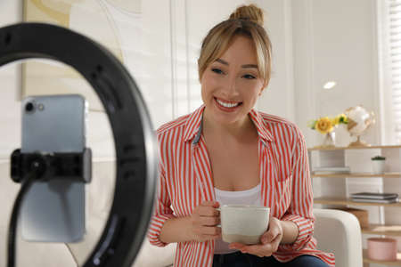 Blogger with cup of tea recording video in living room at home. Using ring lamp and smartphone