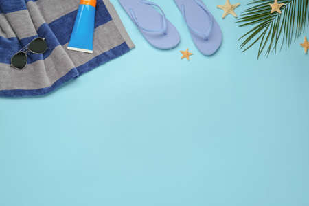 Beach towel, flip flops, sunglasses and sun protection product on light blue background, flat lay....