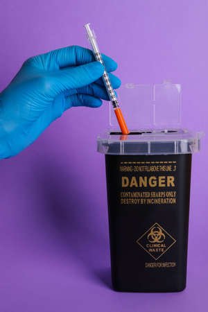 Doctor throwing used syringe into sharps container on violet background, closeup