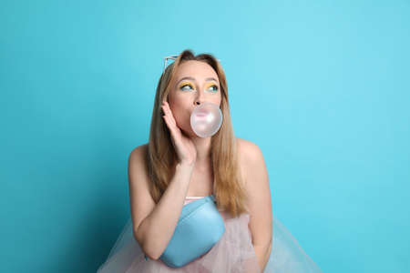 Fashionable young woman with bright makeup blowing bubblegum on light blue background