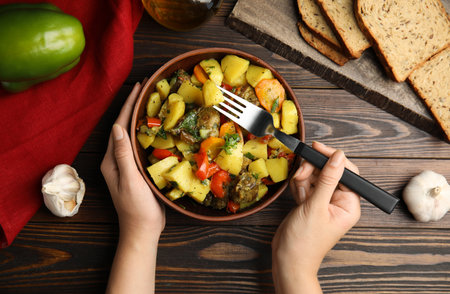 Woman eating delicious dish with potatoes from earthenware at wooden table, top viewの写真素材