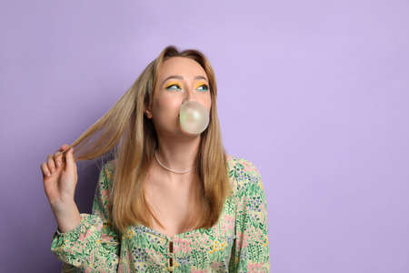 Fashionable young woman with bright makeup blowing bubblegum on lilac background, space for textの写真素材