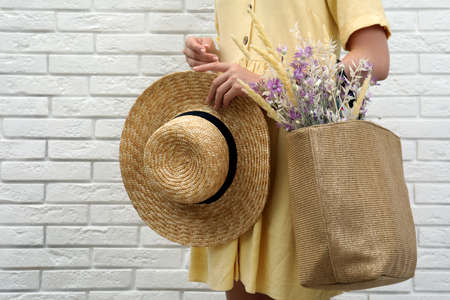 Woman holding straw hat and beach bag with beautiful bouquet of wildflowers near white brick wall, closeupの写真素材