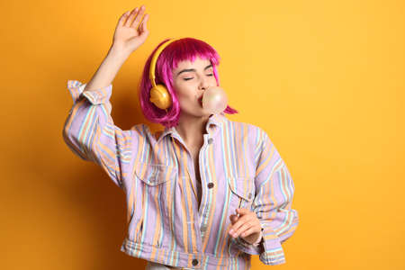 Fashionable young woman in colorful wig with headphones blowing bubblegum on yellow background, space for textの写真素材