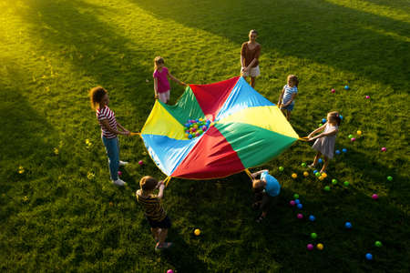 Group of children and teachers playing with rainbow playground parachute on green grass, above view. Summer camp activityの写真素材