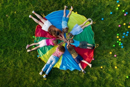 Group of children with teachers holding hands together on rainbow playground parachute in park, top view. Summer camp activityの写真素材