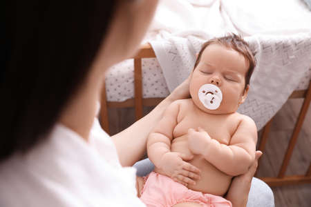 Mother holding her cute little baby with pacifier at home, closeup