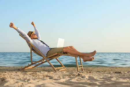Happy man with laptop resting on deckchair near sea. Business tripの写真素材
