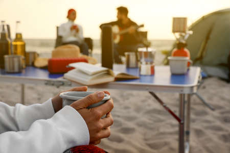 Friends resting near camping tent at beach, focus on woman with cup of hot drinkの写真素材