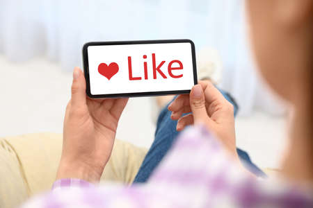 Woman holding smartphone with word Like on screen indoors, closeup