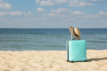 Turquoise suitcase with straw hat on sandy beach near sea, space for textの写真素材
