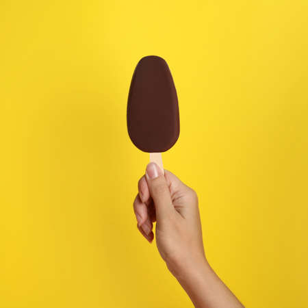 Woman holding ice cream glazed in chocolate on yellow background, closeup