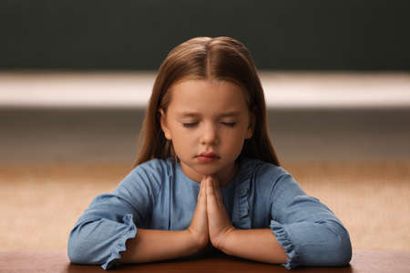 Cute little girl with hands clasped together praying at table indoorsの写真素材