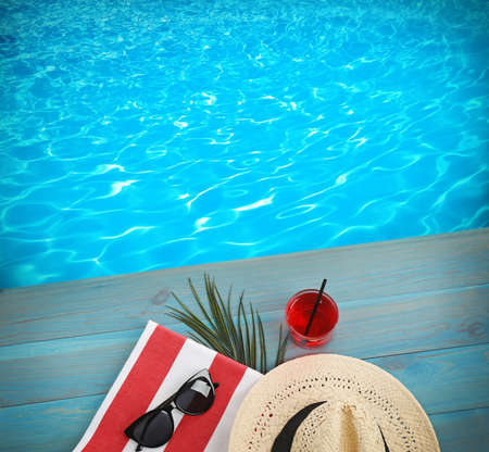 Beach towel, sunglasses, straw hat and refreshing drink on blue wooden surface near swimming pool,...